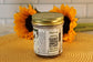 A jar of VGF Cookies and Cream by Sunflour Baking Company with a gold lid sits on a mat; two sunflowers bloom behind it.