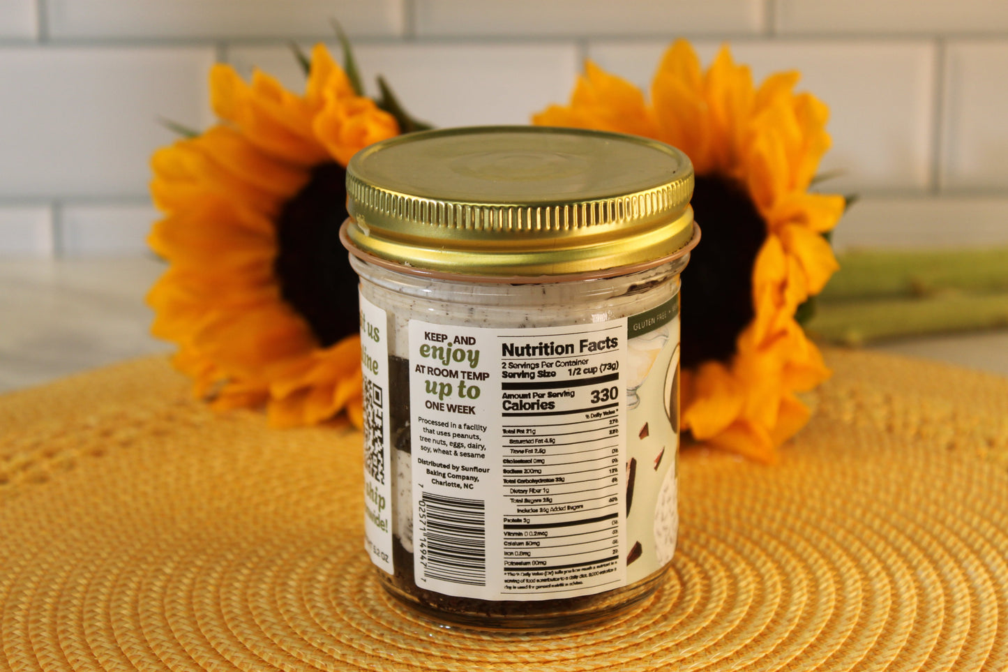 A VGF Cookies and Cream Cake Jar by Sunflour Baking Company sits on a mat, nutrition facts visible, with sunflowers in the background.