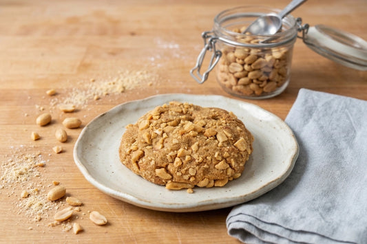 A Sunflour Baking Company Peanut Butter Cookie sits on a plate beside a gray napkin, with peanuts and crumbs on wood.