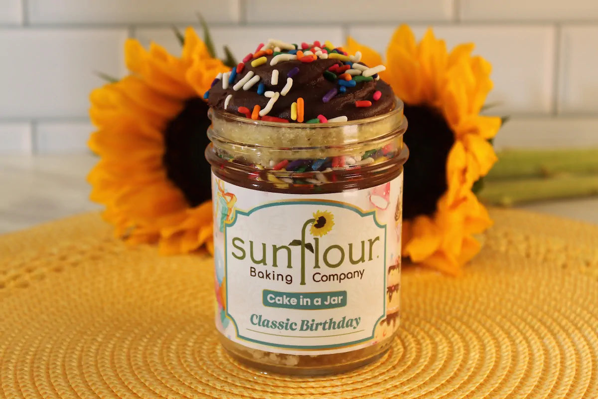 The Sunflour Baking Company Classic Birthday Cake Jar sits on a yellow mat with sunflowers in the background.