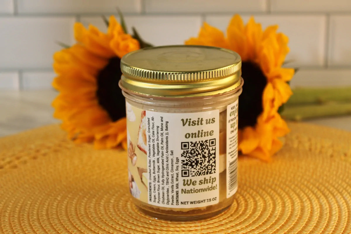 A French Toast Cake Jar by Sunflour Baking Company in glass with a gold lid, labeled with a QR code and shipping info, sunflowers behind.