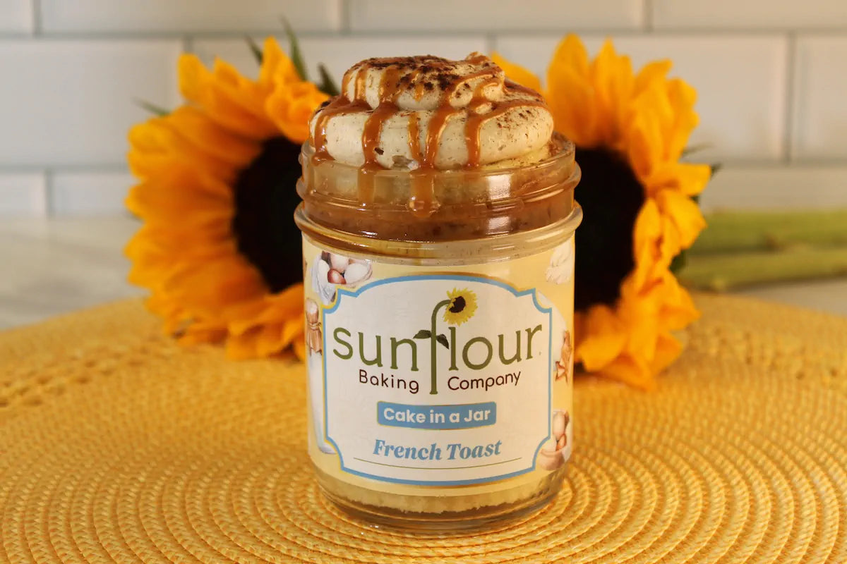 A Sunflour Baking Company French Toast Cake Jar sits on a yellow mat, with two sunflowers behind it.