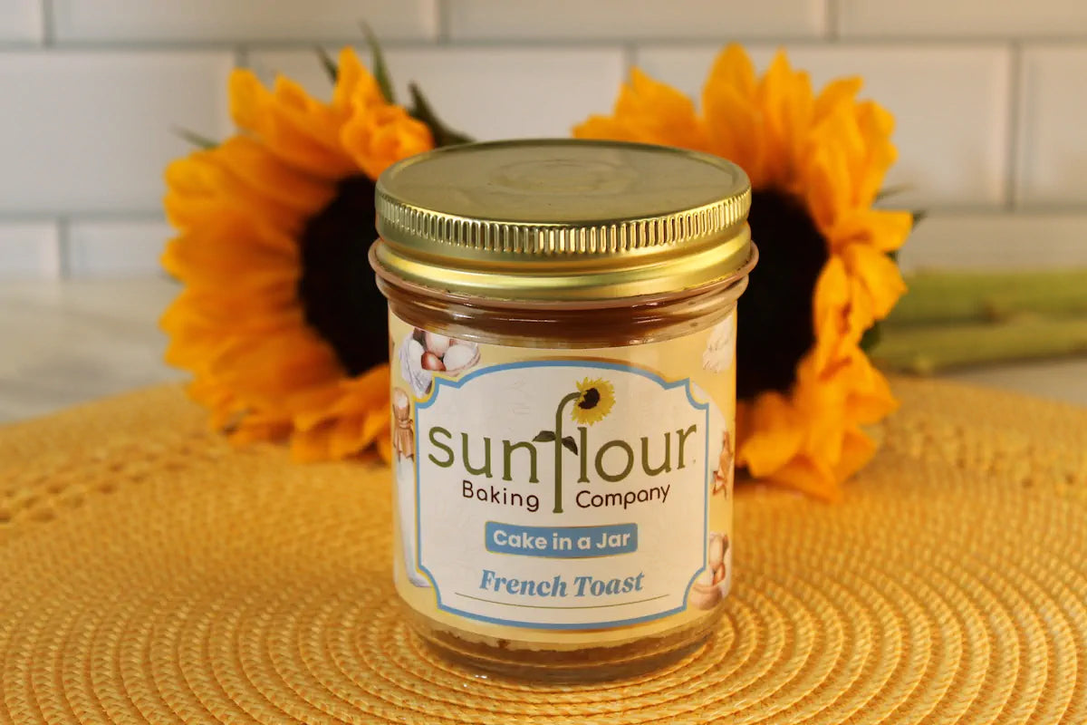 A Sunflour Baking Company French Toast Cake Jar sits on a yellow mat with two sunflowers in the background.